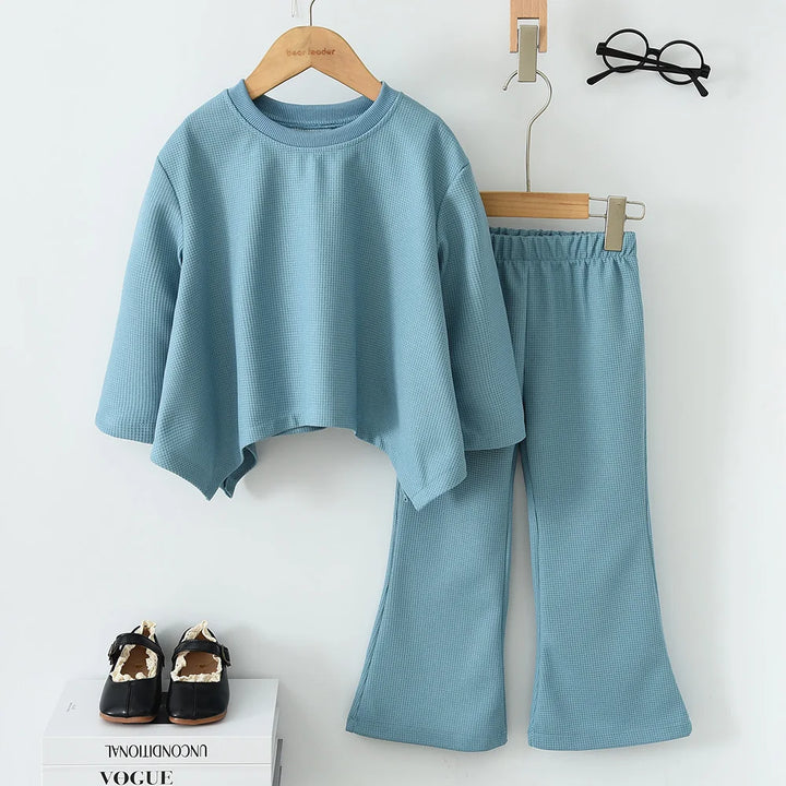 Light blue textured top and pants set on hangers with a magazine and shoes below on a white background